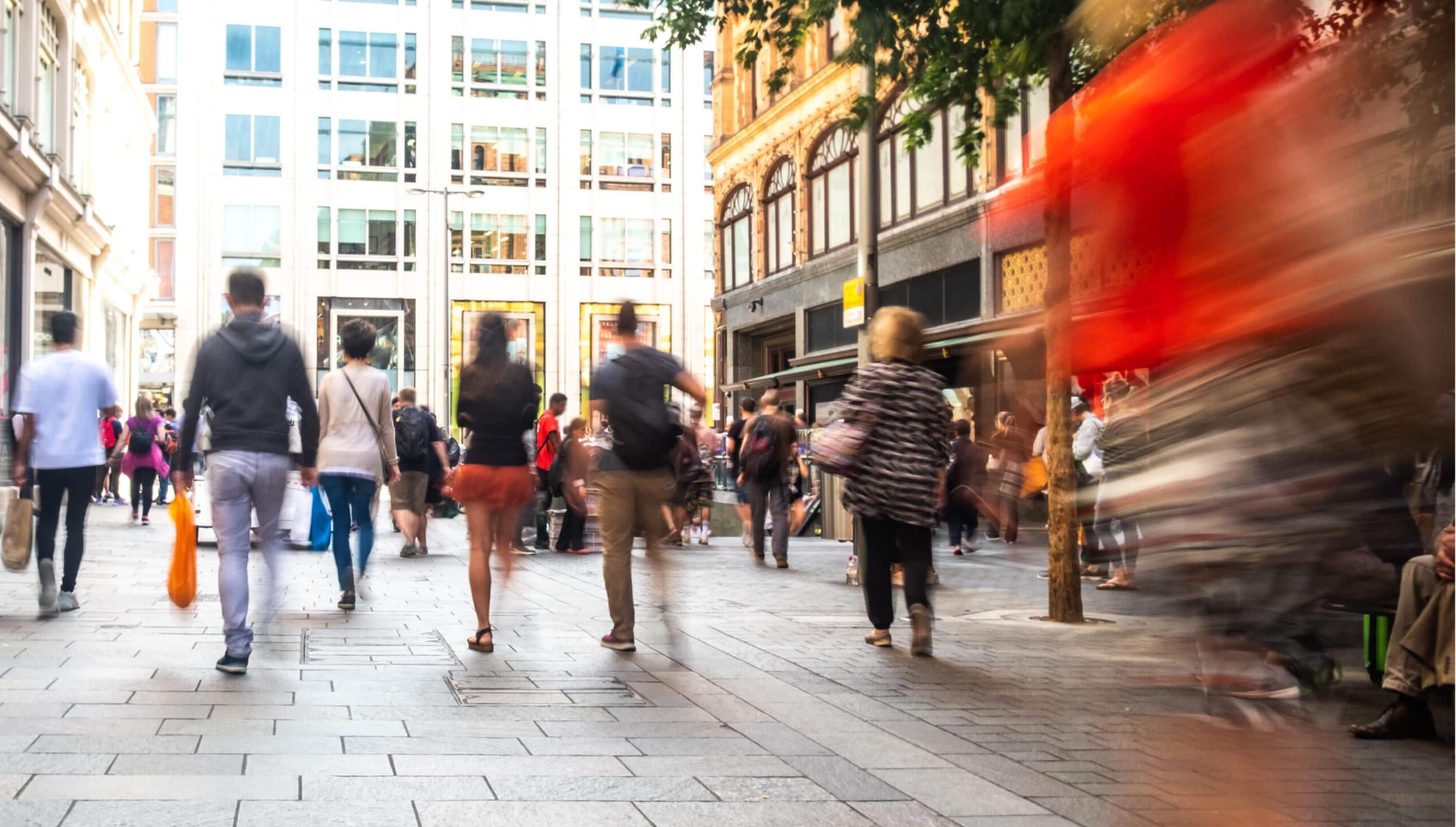 Various people walking in a street with shops either side, some carrying shopping bags. Blue effect on some of the people shown.